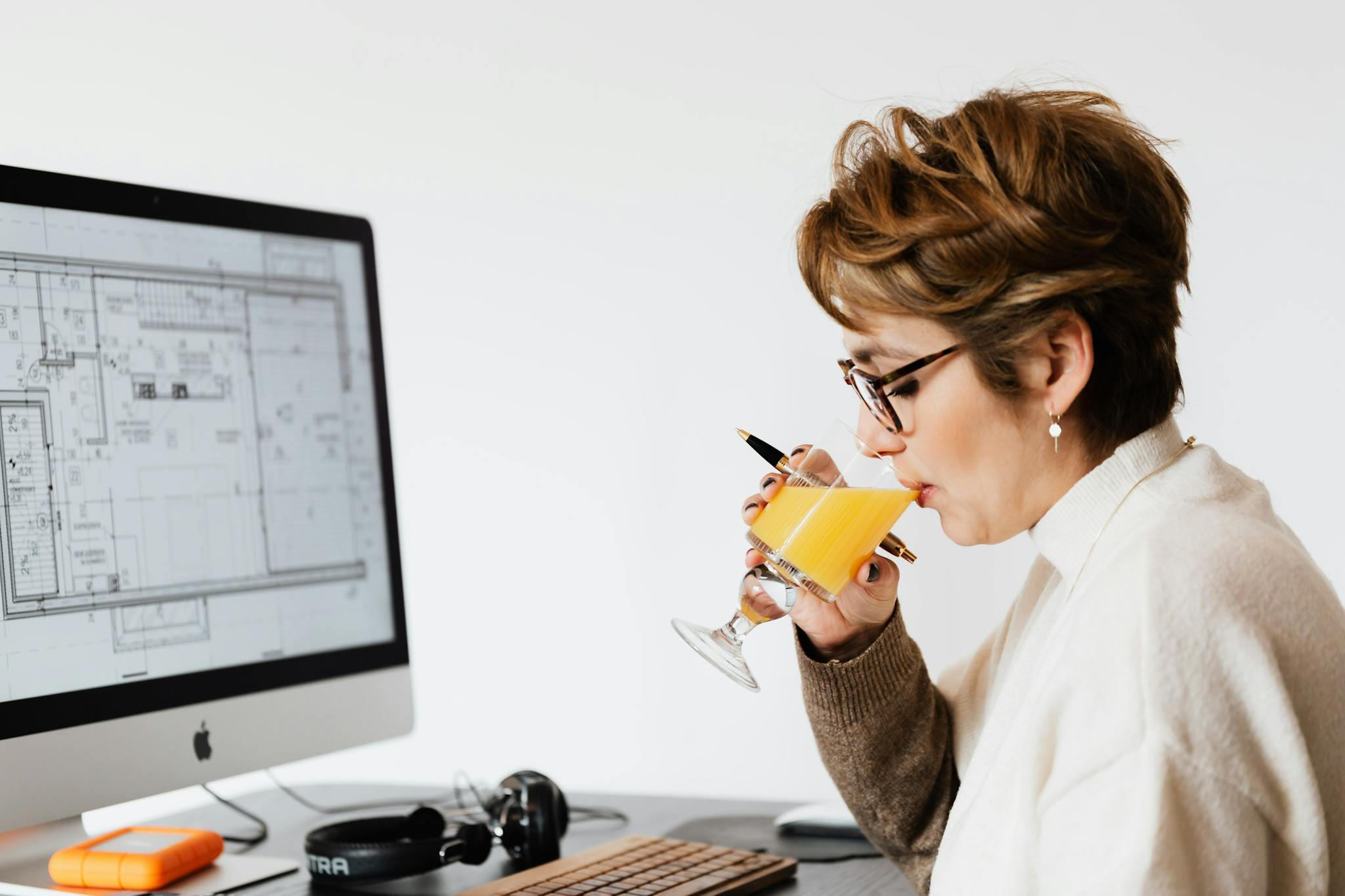 Woman architect working at her desk, focused on computer design plans, drinking juice.